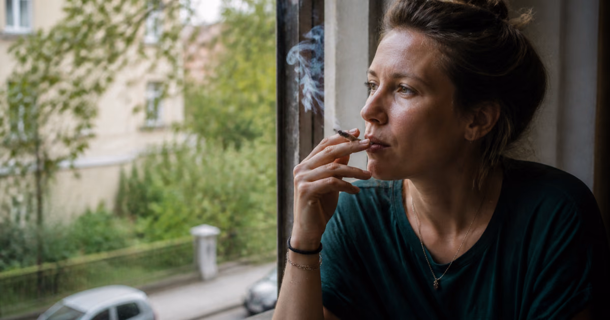 Naine suitsetab akna ääres kanepisigaretti ja vaatab puudega ääristatud tänavale